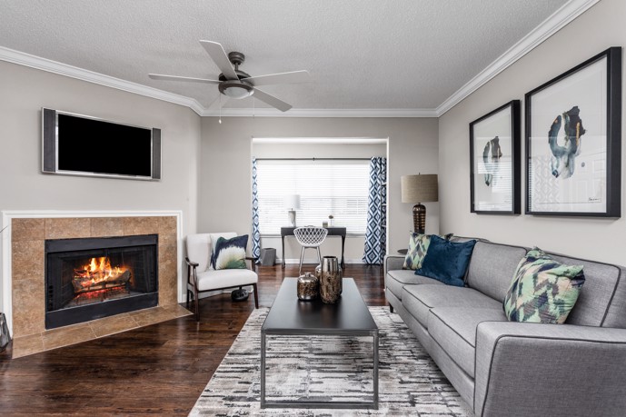 Grey apartment living room furnished with a grey sofa facing a gas fireplace, dark hardwood floors with an area rug, and large artwork and a flat screen tv hanging on the walls