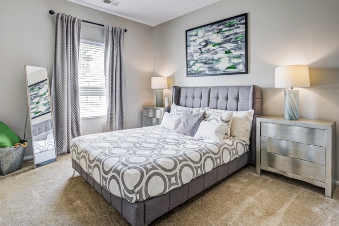 Experience a cozy and comfortable bedroom at The Commons at Canal Winchester, complete with plush carpeting, a queen bed, a window with drapes, and two large side tables to store your belongings.