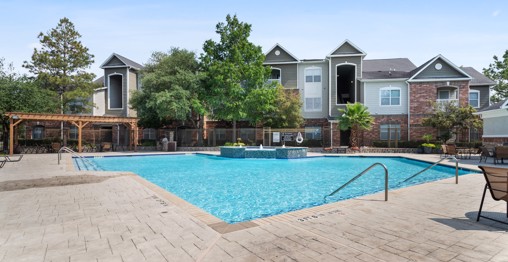 Outdoor community apartment swimming pool with a veranda on the left end and trees and buildings in the background