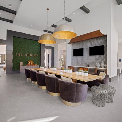 Clubhouse boasting a lengthy community-style wooden table, adorned with contemporary barrel chairs, illuminated by two grand hanging pendant lights.