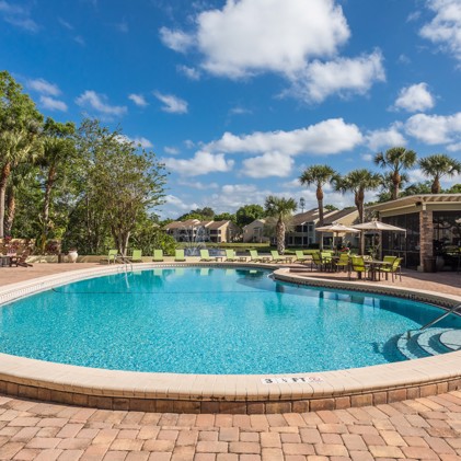 A refreshing swimming pool surrounded by towering trees, lounge chairs, and umbrellas, set against the backdrop of a tranquil pond with a fountain.