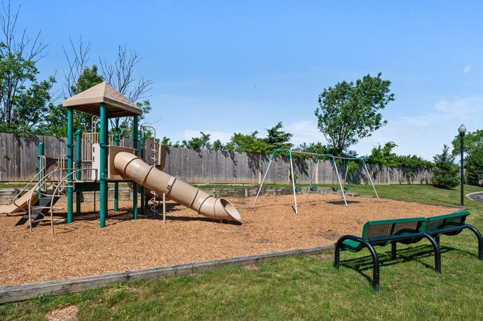 Community outdoor playground on an wood chip ground, surrounded by a lawn and wooden fence with trees in the background