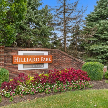 A prominent brick Hilliard Park sign, adorned with flowers, trees, and bushes, serves as a picturesque focal point within the landscape.