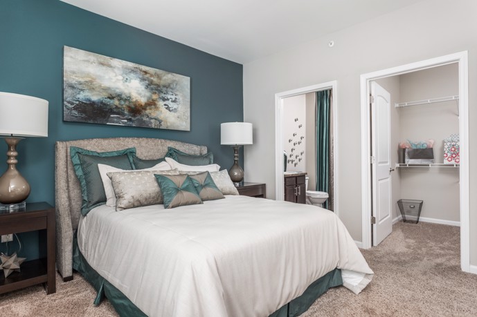 Furnished teal and grey apartment bedroom with a comfortable bed, nightstands with table lamps, an open closet, and door opening to the bathroom on the right