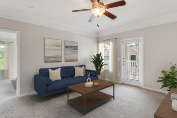 Open-concept grey carpeted apartment with a ceiling fan and a view of the small classic kitchen in the back