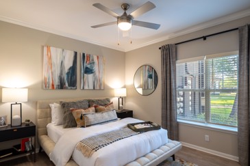 An apartment at The Pointe at Vista Ridge features a bedroom with a queen bed, a ceiling fan, and a large window, offering comfort and natural light.