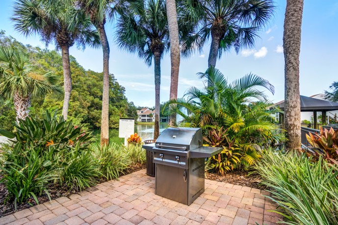 An outdoor grilling area amidst tropical plants and trees near a serene pond at Lakes of Northdale.