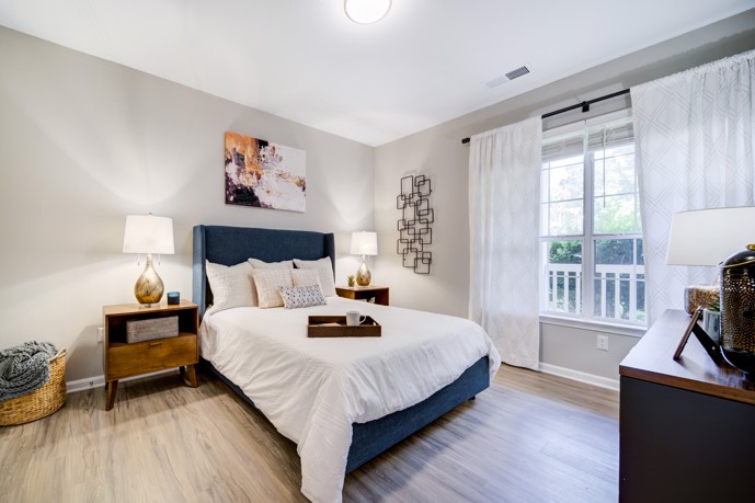 Spacious windows, elegant wood-style flooring, and a plush queen bed adorn the interior of Thornhill apartments in Raleigh, North Carolina, creating a serene and inviting atmosphere for residents.