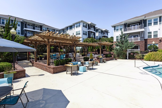 Outdoor community seating area that is paved and has brown wooden gazebo next to the swimming pool and apartment buildings in the background