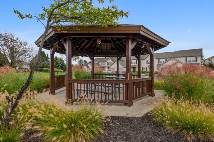 A charming gazebo, furnished with two picnic tables, nestled amidst vibrant plants, trees, and grass.