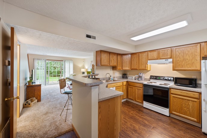A spacious U-shaped kitchen boasting hardwood-style flooring, a breakfast bar, and wooden cabinets, seamlessly connected to a cozy carpeted living room.