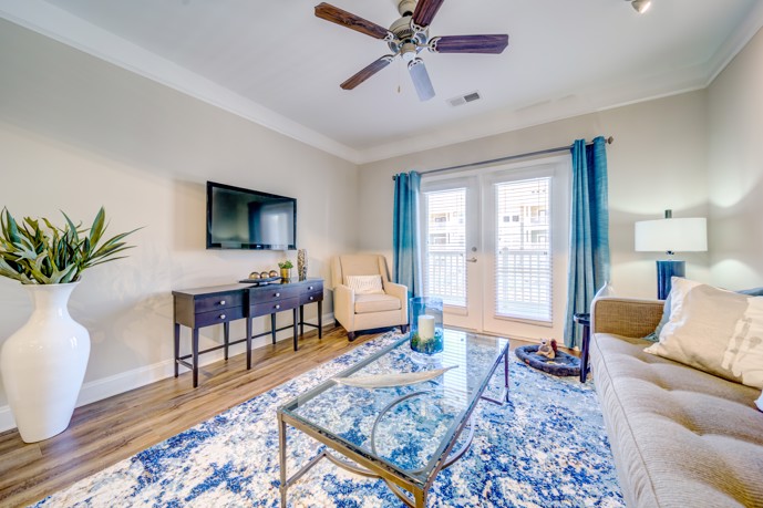 Comfortably furnished apartment living room with hardwood flooring, flat screen tv, a ceiling fan, beige seating, and blue and white drapes and area rug