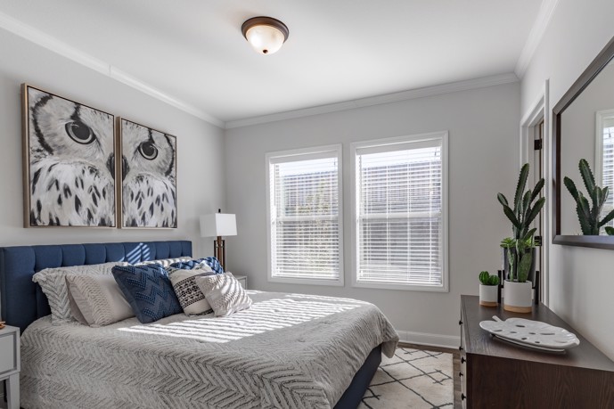 A queen bed in a carpeted bedroom with two sizable windows and a prominent owl painting at Virtuoso Apartments.