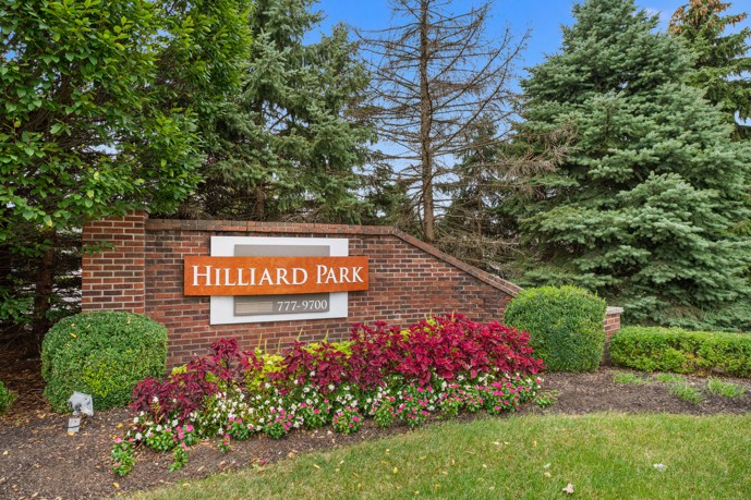 A prominent brick Hilliard Park sign, adorned with flowers, trees, and bushes, serves as a picturesque focal point within the landscape.