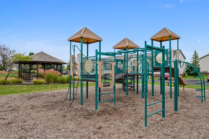 A sprawling playground featuring wood chips, positioned conveniently near a gazebo, a parking lot, and expansive green spaces.
