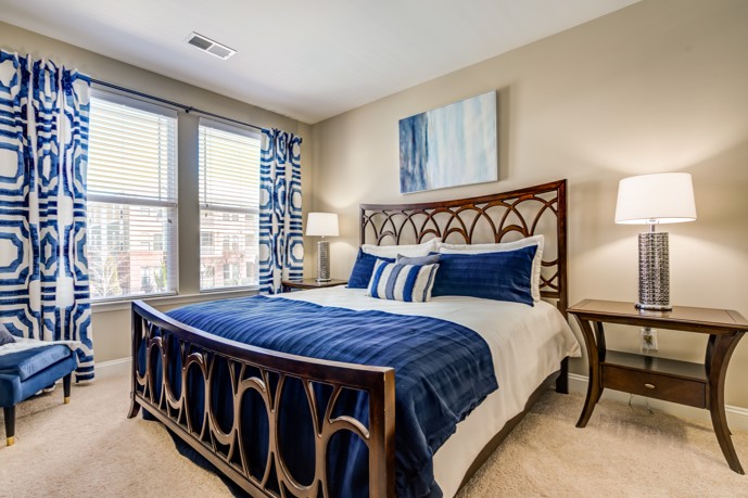 Serene apartment bedroom furnished with tan wall-to-wall carpeting, nightstands with table lamps, and blue and white drapes and bedding