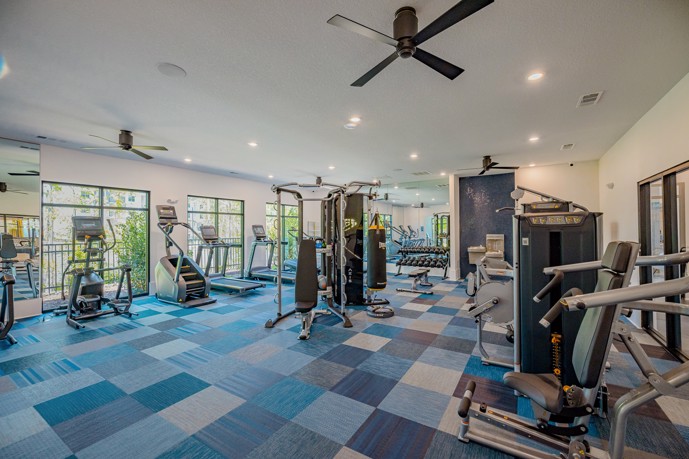 Apartment community fitness center with blue-squared tiled carpeting, ceiling fans,  and exercise machines along the perimeter and down the center of the room