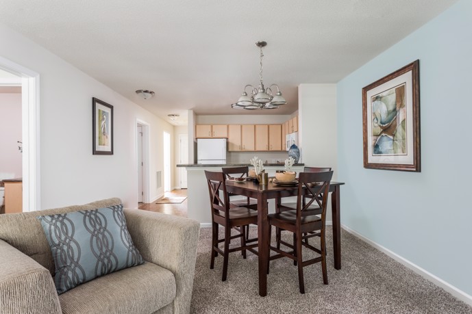 A cozy carpeted open-plan living room, seamlessly connected to a dining area and kitchen, providing residents with a spacious and inviting living space at The Tides of Calabash in Sunset Beach, NC.