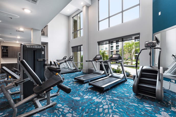 Blue and white indoor apartment community fitness center with exercise equipment facing windows