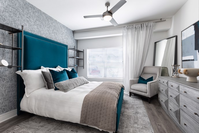 Grey apartment bedroom furnished with a blue bed, grey armchair, credenza, mirrors, a large window, and a ceiling fan
