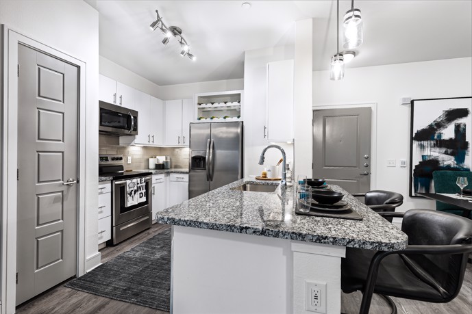 An apartment kitchen with white walls and cabinets, grey granite countertops, stainless steel appliances, and an island with black stools facing it