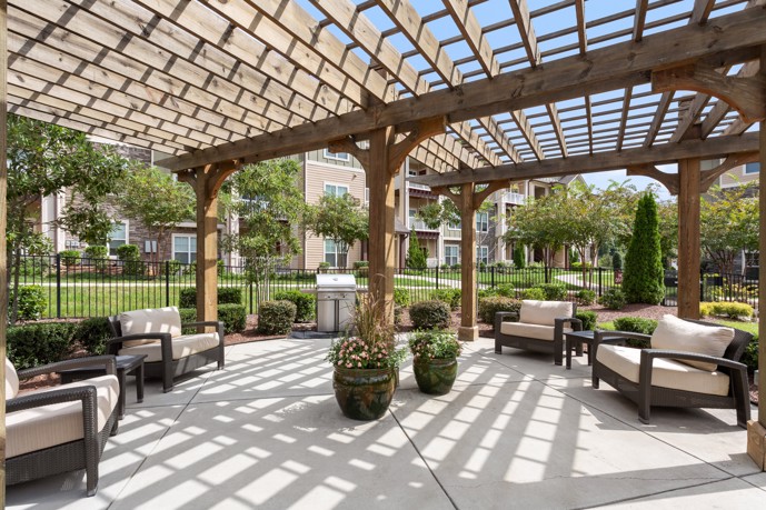 Sunny exterior view of a community picnic area under a wooden pergola near the grilling station with apartments, trees, and an iron fence in the background