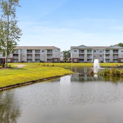 A tranquil lake adorned with a fountain, creating a scenic focal point in front of the apartments at The Tides of Calabash, in Sunset Beach, NC, enhancing the natural beauty of the surroundings.