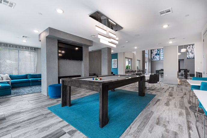Blue and grey apartment clubhouse with a grey pool table in the middle, a seating area to the left, and tables and chairs to the right and in the back