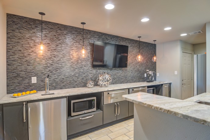 Community clubhouse modern kitchen with grey cabinets, tile backsplash with a flat screen TV, granite countertops, and microwaves