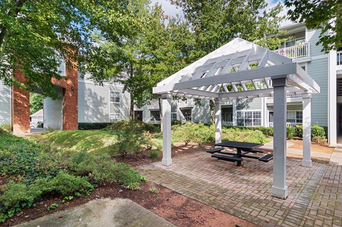 Outdoor gazebo adorned with a picnic table, nestled amidst verdant plants, lush grass, and towering trees within the tranquil grounds of Jefferson at the Perimeter.