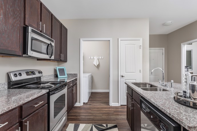 Apartment kitchen with dark wood cabinets, granite countertops, and stainless steel appliances with a view facing the small laundry room