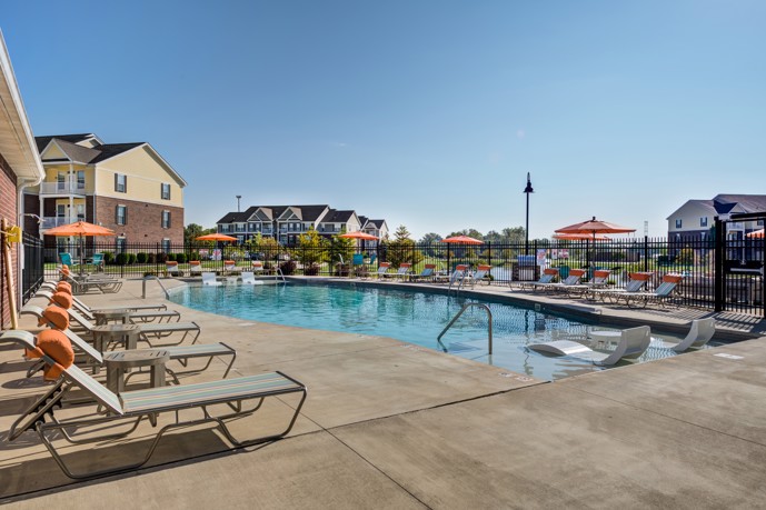 Outdoor community swimming pool on a sunny day with lounge chairs lined along the left side and an iron fence and apartments behind it in the background