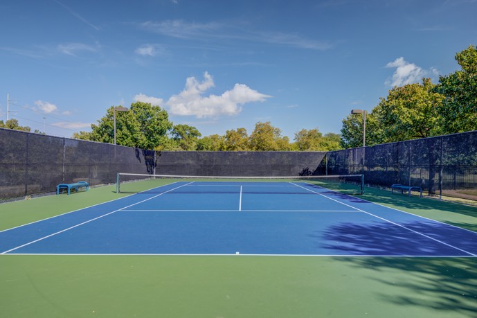 A meticulously maintained tennis court, generously sized and accompanied by a convenient bench, set amidst the open green space.