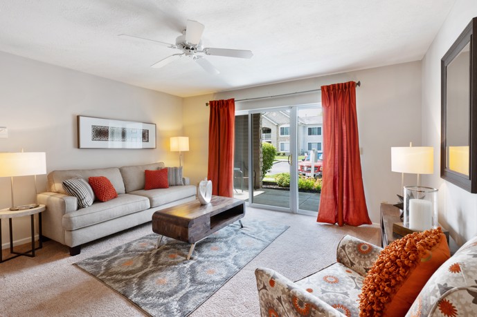 Comfortably furnished apartment living area with wall-to-wall carpeting, a sofa and armchair with rust-colored throw pillows that match the drapes over a sliding glass door 