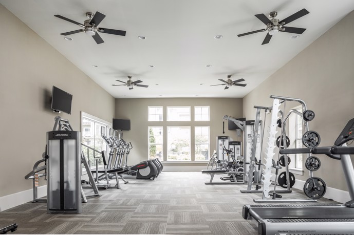 Long room view of a community apartment fitness center facing windows with exercise equipment along the left and right walls and four ceiling fans