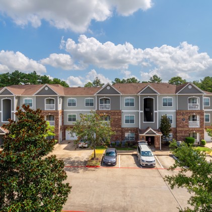 Front aerial view of the parking lot with two cars and the Carrington Park at Huffmeister apartment buildings in Cypress, TX behind it