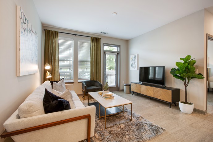 Experience a modern and inviting apartment at Vesta City Park, complete with light wood-style flooring, a comfortable large two-seater couch, and large windows that provide ample natural light.