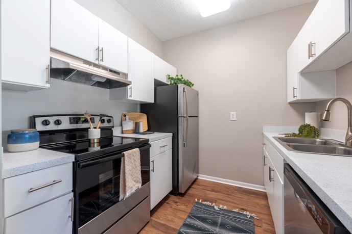 Long view of a small tan apartment kitchen with white cabinets, stainless steel appliances and a sink 