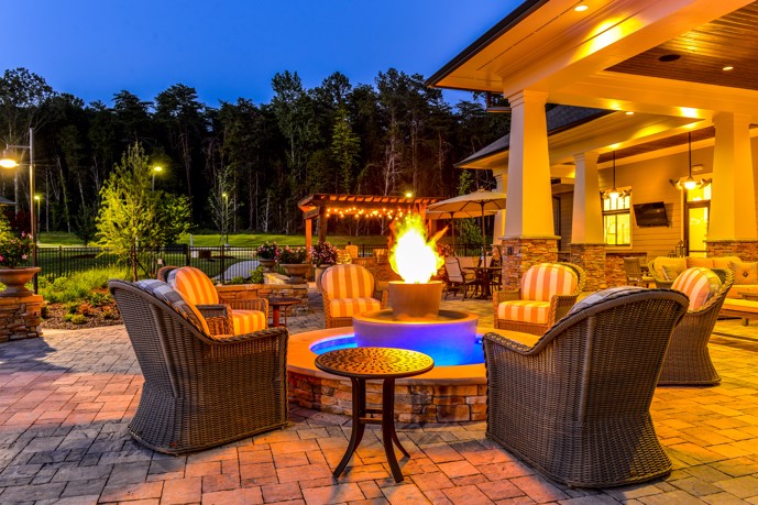 Outdoor fireside lounge equipped with cozy chairs, a fire pit, tables adorned with umbrellas, and string lights hanging under a pergola.
