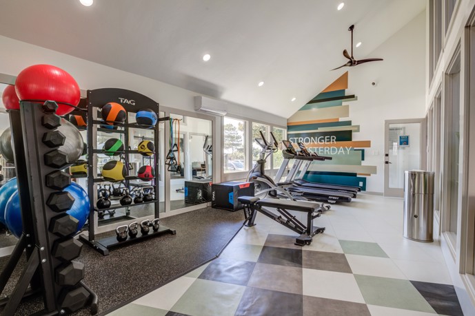 The fitness center within The Invitational apartments, equipped with a variety of gym equipment, enhanced by ample natural light streaming in through large windows and mirrored walls, creating an energizing atmosphere for workouts.