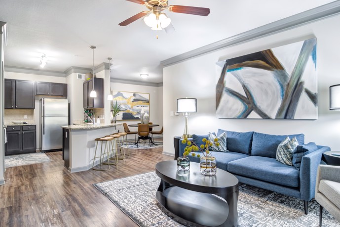 At Waterstone at Big Creek, discover an open floor plan apartment adorned with a stylish blue two-seater couch, a kitchen complete with a bar, and a dining area featuring a table.